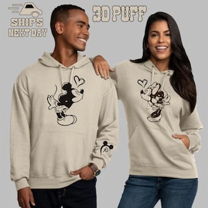 Könnte beinhalten: Ein Paar trägt passende beige Hoodies mit einem 3D-Puffdruck von Mickey Mouse und Minnie Mouse, die sich küssen. Der Text "3D Puff" ist oben im Bild gedruckt. Der Mann trägt einen Hoodie mit Mickey Mouse auf der Vorderseite und einem Mickey Mouse-Ohr am Ärmel. Die Frau trägt einen Hoodie mit Minnie Mouse auf der Vorderseite.