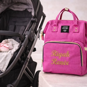 Può includere: Una borsa per pannolini rosa acceso con dettagli dorati e il nome "Riyah Renée" ricamato in oro. La borsa ha un manico superiore e una tasca frontale. Un passeggino e scarpine rosa sono sullo sfondo.