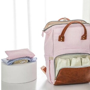 May include: A pink and white striped diaper bag with brown leather accents. The bag has multiple pockets and a zippered closure. The bag is open and the inside is lined with a light beige fabric.