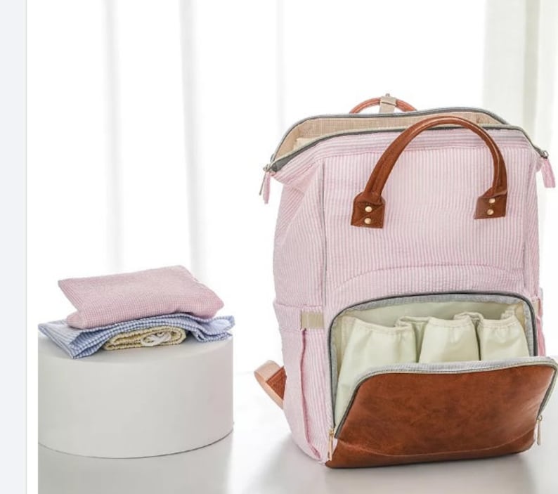 May include: A pink and white striped diaper bag with brown leather accents. The bag has multiple pockets and a zippered closure. The bag is open and the inside is lined with a light beige fabric.