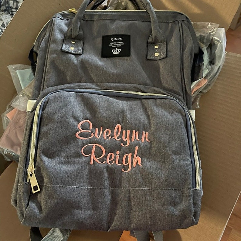 Gray canvas diaper bag with a zippered front pocket and a gold zipper pull.  The front pocket has the name "Evelyn Reigh" embroidered in coral.