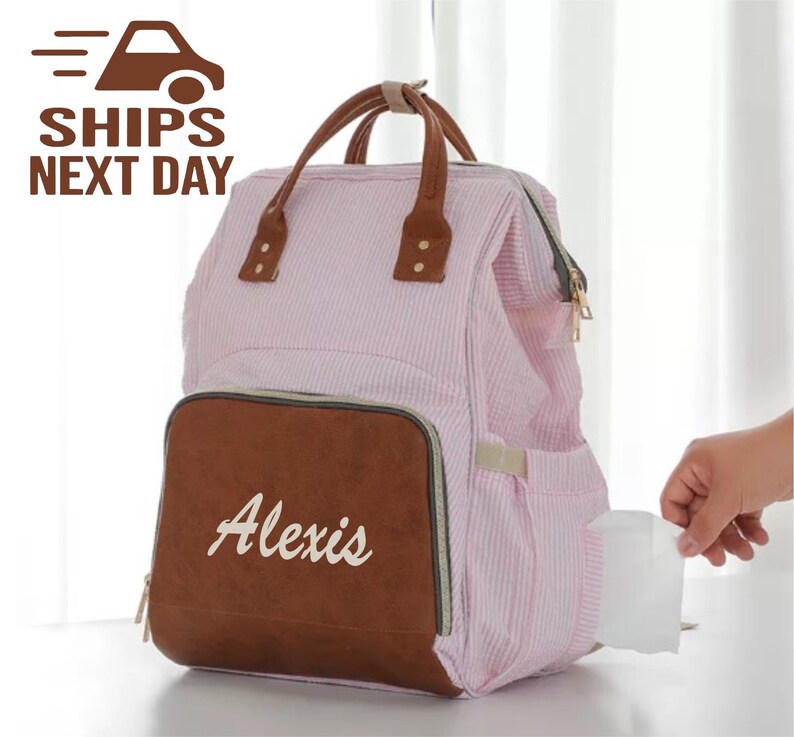 May include: Pink and brown striped diaper bag backpack with a brown leather flap that says "Alexis" in white script. The bag has a zippered pocket on the side.
