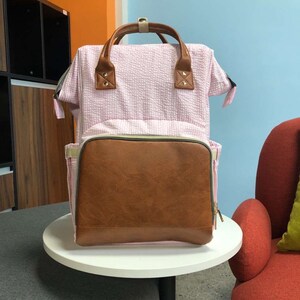 May include: A pink and brown diaper bag with a quilted exterior and a faux leather front pocket. The bag has adjustable straps and a top handle.
