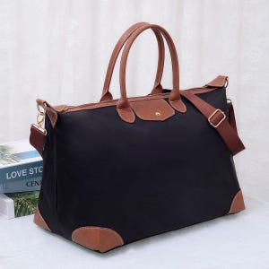 May include: Black travel tote bag with brown leather detailing. Features include a top handle, shoulder strap, and leather corner and flap details. The bag has a rectangular shape and a gold-coloured clasp.