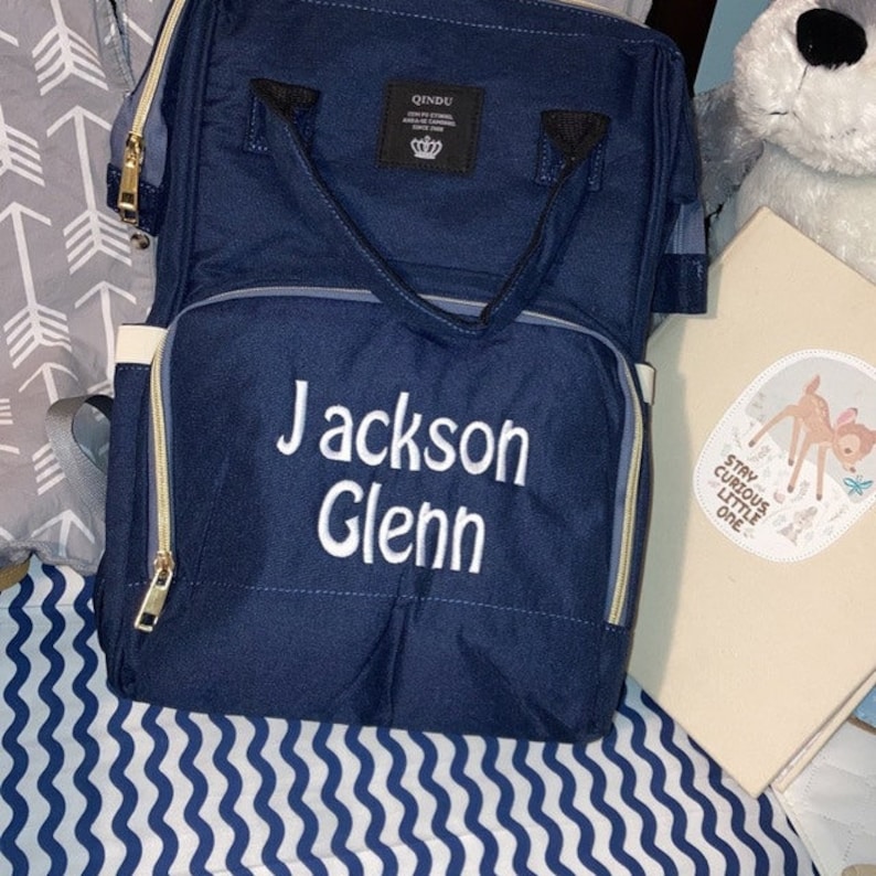 A navy blue diaper bag with a white zipper and a white embroidered name "Jackson Glenn". The bag has a top handle and adjustable straps. The bag is sitting on a white and blue striped surface.