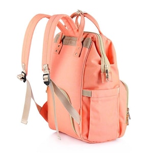 K&ouml;nnte beinhalten: Ein peachfarbener Wickeltasche-Rucksack mit mehreren Taschen und verstellbaren Tr&auml;gern. Die Tasche hat eine Rei&szlig;verschlusstasche vorne und ein gro&szlig;es Hauptfach.