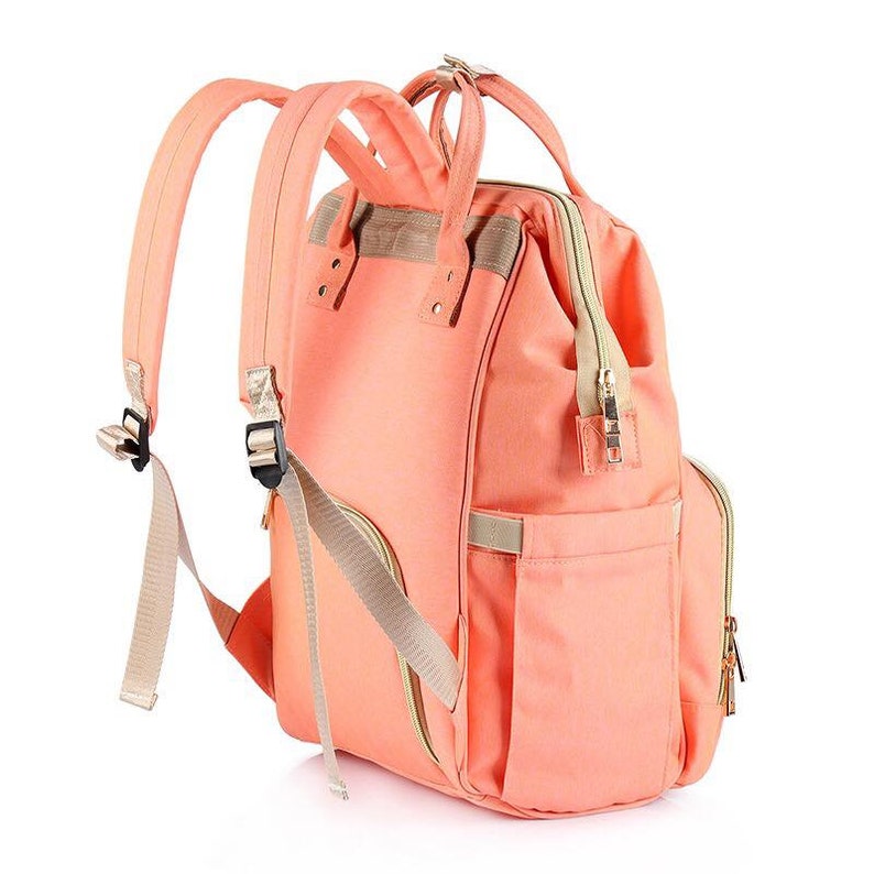 K&ouml;nnte beinhalten: Ein peachfarbener Wickeltasche-Rucksack mit mehreren Taschen und verstellbaren Tr&auml;gern. Die Tasche hat eine Rei&szlig;verschlusstasche vorne und ein gro&szlig;es Hauptfach.