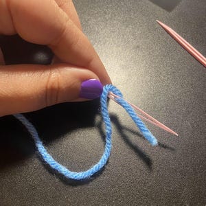 May include: Close-up of a light blue yarn loop being worked with a pink knitting needle. The needle is inserted through the yarn, creating a stitch. The background is a dark surface.