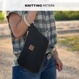 May include: A black knitted pouch with a zipper closure and a wooden handle. The pouch is worn over the shoulder of a person wearing a plaid shirt and blue jeans.