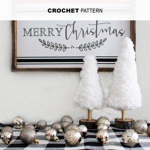 May include: A white and black checkered table runner with silver Christmas ornaments and two white fluffy Christmas trees. A wooden sign with the text "Merry Christmas" hangs on the wall behind the table runner.