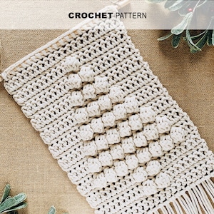 Puede incluir: Un tapiz de pared de crochet blanco con un patrón geométrico y flecos. El tapiz está hecho con un hilo grueso y tiene un estilo rústico y bohemio.