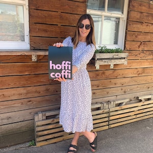 Könnte beinhalten: Eine Frau in einem weißen Kleid mit schwarzen Punkten hält ein schwarzes Schild mit den Worten "hoffi coffi" in rosa Buchstaben.