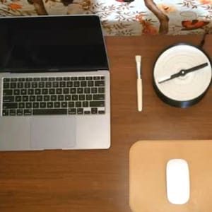 May include: A laptop computer with a black screen and a silver keyboard is on a wooden desk. A small, round, white and black object with a propeller is on the desk. A brown leather mouse pad with a white mouse is on the desk. A wooden paintbrush is on the desk.