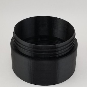 May include: A black plastic container with a smooth, cylindrical shape and a slightly raised rim. The container has a textured surface and appears to be made from 3D printed plastic.