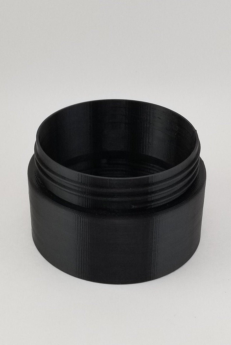 May include: A black plastic container with a smooth, cylindrical shape and a slightly raised rim. The container has a textured surface and appears to be made from 3D printed plastic.