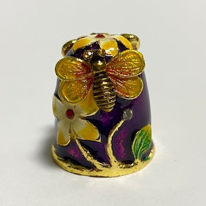 Puede incluir: Un dedal decorativo con base morada, adornado con detalles dorados y una mariposa. El dedal presenta flores amarillas y blancas con centros rojos. La mariposa tiene alas amarillas y cuerpo dorado. Una pequeña joya está engastada en el lateral.