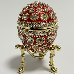 Puede incluir: Un huevo rojo adornado con joyas sobre un soporte dorado. El huevo está decorado con diamantes de imitación transparentes y tiene una banda dorada en el medio. El soporte tiene tres patas y también está decorado con diamantes de imitación.