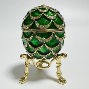 Könnte beinhalten: Eine dekorative, eiförmige Schmuckschatulle aus grünem Emaille mit goldenen Akzenten und Kristallverzierungen. Das Ei steht auf einem goldfarbenen Ständer mit klauenartigen Füßen. Die Oberfläche des Eis hat ein strukturiertes, geschupptes Design.