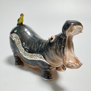 May include: A decorative hippo figurine with an open mouth, painted in shades of gray, brown, and beige. A small yellow bird sits on its back. The hippo has a jeweled accent around its body.