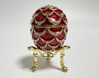 May include: A decorative red egg-shaped trinket box with a gold-colored stand. The egg has a textured, scaled design with gold trim and small, clear gemstones. The stand has a detailed, ornate design.
