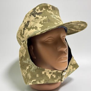 Puede incluir: Un sombrero de camuflaje con ala y cubrecuello, exhibido en una cabeza de maniquí. El sombrero presenta un diseño digital pixelado en tonos verdes, beige y marrón. El cubrecuello se asegura con dos botones.