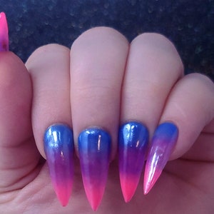May include: A hand displaying long, pointed artificial nails. The nails feature a gradient design, transitioning from blue at the base to pink at the tips. The nails have a glossy finish.