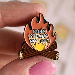 May include: Enamel pin featuring a campfire design with orange flames and brown logs. The pin has the text "BURN STEADY, NOT OUT" in a white and yellow font. The pin is held by a person.
