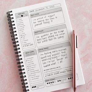 a spiral-bound notebook with a pen resting on top of it, displaying a self-care journal page with prompts for reflection and self-care activities.