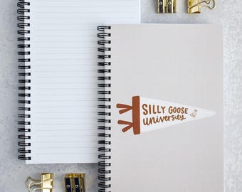 May include: Two spiral-bound notebooks with black wire binding. One notebook is open to lined white pages, while the other is closed with a light gray cover featuring a brown pennant graphic that reads Silly Goose University. Two gold metal clips and two pencil sharpeners are included.