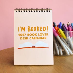May include: A spiral-bound desk calendar with a light beige cover featuring the orange text I'M BOOKED! 2027 BOOK LOVER DESK CALENDAR and a graphic of an open book with white pages and an orange spine, positioned next to a clear cup of markers.