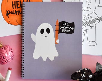 May include: A spiral-bound coloring book with a gray cover featuring a white ghost illustration holding an orange crayon and a black flag that reads "FALL COLORING BOOK". Other coloring pages and markers are visible in the background.