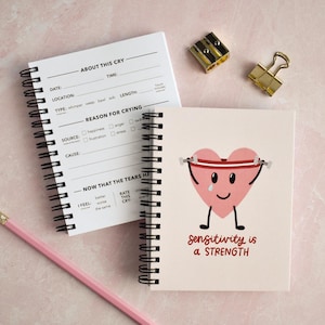 May include: Two spiral-bound notebooks with lined pages and a pink pencil. One notebook has fill-in-the-blank prompts about crying. The other notebook features a pink heart character with the text "sensitivity is a STRENGTH". A gold paper clip and pencil sharpener are also visible.