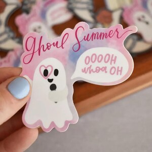 May include: A die-cut sticker featuring a white ghost character holding a microphone. The ghost has a pink heart-shaped eye and is singing. The text reads Ghoul Summer and OOOOH whoa OH in pink script and block letters against a watercolor background.