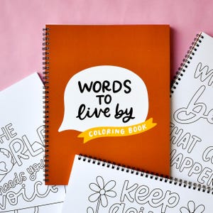 May include: An orange spiral-bound coloring book titled "WORDS TO live by COLORING BOOK" in a white speech bubble. Several other coloring books with inspirational quotes are visible, all on a pink background.