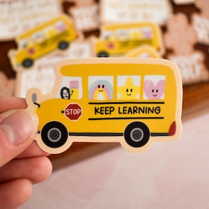 May include: A yellow school bus sticker featuring a red stop sign, black wheels, and windows displaying a pencil, a rainbow, a yellow star, and a pink heart. The text KEEP LEARNING is printed in black on the side of the bus body.