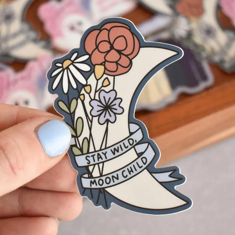 May include: A crescent moon sticker featuring a bouquet of flowers including a large coral bloom, a white daisy, and a small purple flower. A banner wrapped around the moon displays the text STAY WILD, MOON CHILD in black capital letters.