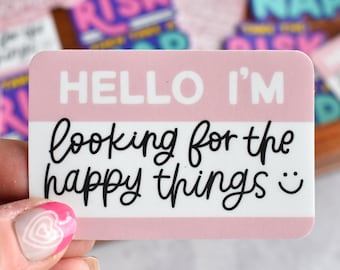 Puede incluir: Una pegatina rectangular con una parte superior rosa claro y una inferior blanca. La parte superior presenta el texto blanco HELLO I'M. La parte inferior contiene el texto cursivo negro looking for the happy things y una cara sonriente negra.