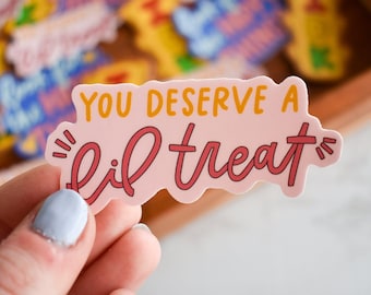 May include: A die-cut sticker with a light pink background featuring the orange text YOU DESERVE A and the dark pink cursive text lil treat. The sticker has a wavy border and small dark pink accent lines on the left and right sides of the text.