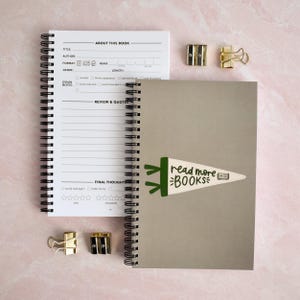 May include: Two spiral-bound notebooks with black wire binding. One notebook is open, showing a printed page with sections for book title, author, and reviews. The other notebook is closed with a beige cover featuring a green pennant graphic with the text read more BOOKS and a small book icon. Two gold metal pencil sharpeners and two gold metal binder clips are included.