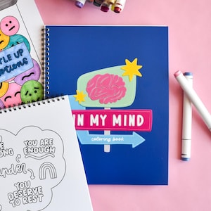 May include: A blue spiral-bound coloring book with the words "IN MY MIND" and a brain graphic. The cover also features a signpost design with a star and arrow. Other coloring books and markers are also visible.