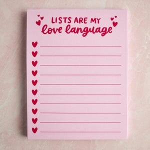 May include: A pink notepad with the phrase "LISTS ARE MY love language" in red script. Red heart illustrations are scattered around the text and along the left side. The notepad has horizontal lines for writing.