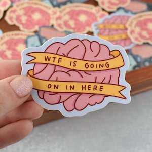 May include: A die-cut sticker featuring a pink illustration of a human brain wrapped in a yellow ribbon. The ribbon contains the black text "WTF IS GOING ON IN HERE" written in a simple, sans-serif font across two separate horizontal lines.