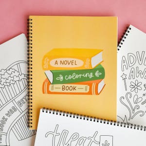 May include: A spiral-bound notebook with a yellow cover featuring a stack of illustrated books labeled "A Novel," "coloring," and "Book." Surrounding the main notebook are other notebooks with black and white line art.