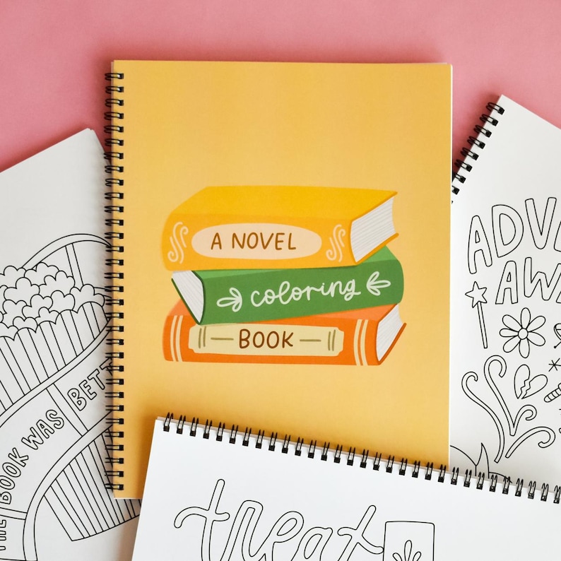 May include: A spiral-bound notebook with a yellow cover featuring a stack of illustrated books labeled "A Novel," "coloring," and "Book." Surrounding the main notebook are other notebooks with black and white line art.