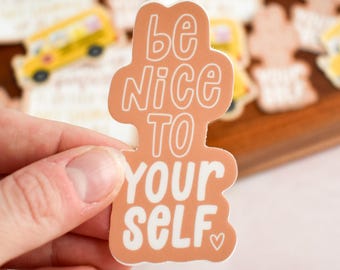 Puede incluir: Una pegatina troquelada con fondo naranja apagado y letras blancas que dicen "be Nice TO YOUR SELF" seguido de un pequeño símbolo de corazón. La pegatina tiene un borde ondulado e irregular y presenta un acabado mate con tipografía negrita.