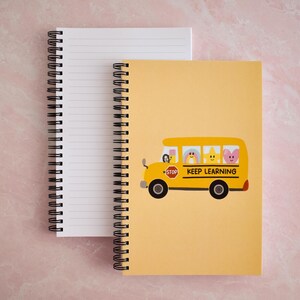 May include: A spiral-bound notebook with a yellow cover featuring a cartoon school bus illustration. The bus has a red stop sign and the text KEEP LEARNING. Inside the bus are a pencil, rainbow, star, and heart. The notebook has white lined pages.