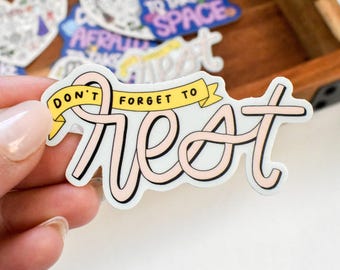 Puede incluir: Una pegatina de vinilo troquelada que presenta el texto "DON'T FORGET TO" en letras negras sobre una cinta amarilla, con la palabra "REST" escrita en letra cursiva grande de color rosa claro con un borde negro sobre fondo blanco.