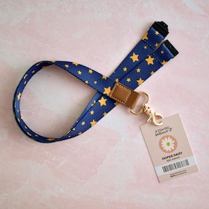 May include: A navy blue lanyard featuring a repeating pattern of small yellow stars. The lanyard includes a brown faux leather connector and a gold-toned metal swivel lobster clasp. Attached is a rectangular tag with the text KYN YOU believe it, OOPSIE DAISY, and a barcode.