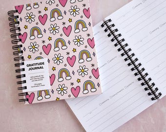 May include: A spiral-bound gratitude journal with a light pink cover featuring a repeating pattern of rainbows, white daisies, pink hearts, and yellow stars. The cover includes a white label with the text GRATITUDE JOURNAL, daily gratitude log with coloring pages and 366 entries, 126 pages, handmade in Texas. The open journal shows lined pages with checkboxes.