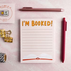 May include: A flat lay featuring stationery and office supplies. A notepad with the text "I'M BOOKED!" and an illustration of an open book. A pink pen, a rose gold pen, a library card pin, and gold binder clips are also visible.